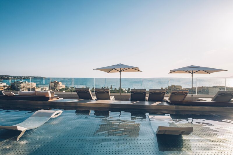 Dachterrasse mit Infinity-Pool und modernen Liegestühlen, einige davon direkt im Wasser. Drei große Sonnenschirme spenden Schatten. Im Hintergrund erstreckt sich ein weiter Blick über das Meer bis zum Horizont.